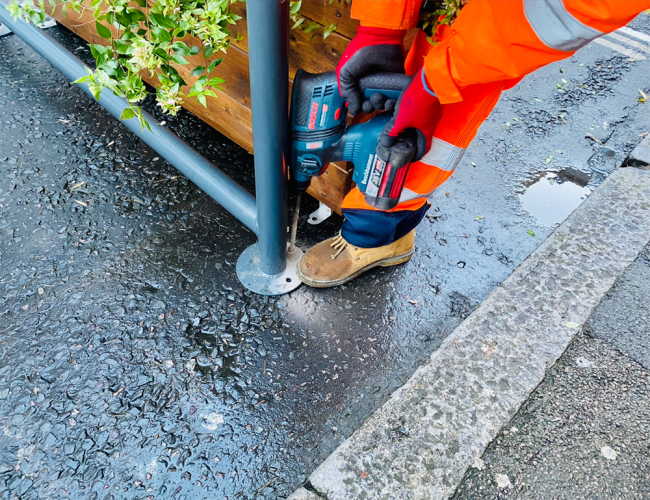 Worker wearing a high-vis vest installs the Mobility Corral into the road using a drill