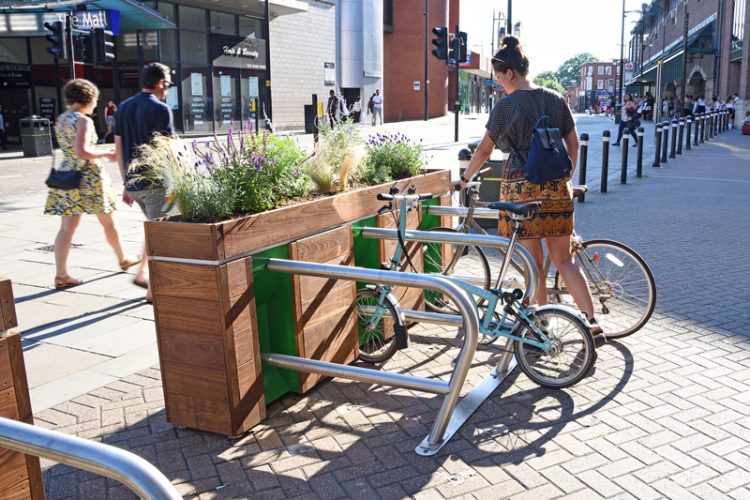 Next to a pedestrianised street a cyclist parking their bike in black stands attached to the Cyclehoop Planter Rack