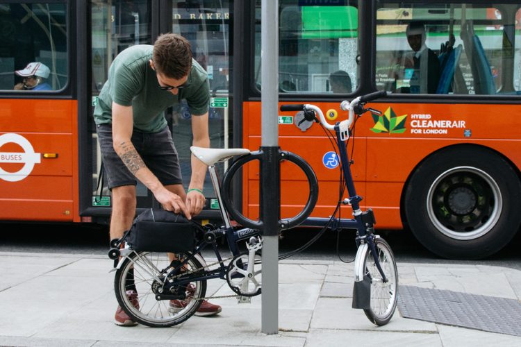 A cyclist locking a foldable bike to the Cyclehoop bike stand on a street as a bus passes by